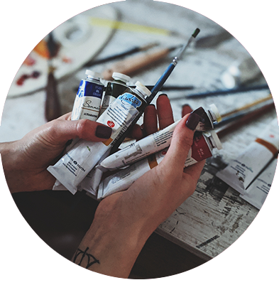 A person holding various paint tubes and a brushes in their hand at their studio workbench.